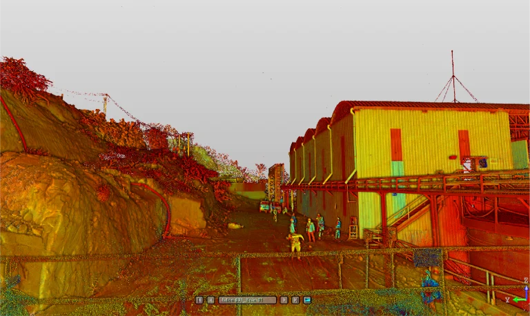 Nube de puntos de Mina DIABRAS muestra a personas cerca de un gran edificio de paredes metálicas, una pasarela de madera y una ladera rocosa a la izquierda. Un vehículo está aparcado más atrás en el camino de tierra