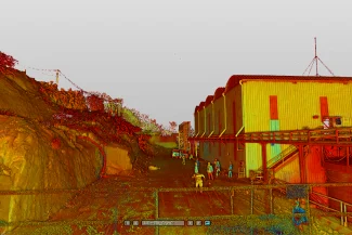 Una nube de puntos digital muestra a un grupo de personas cerca de los edificios industriales de Mina DIABRAS, con una ladera rocosa a la izquierda y un cielo despejado. La escena está coloreada en rojos, amarillos y verdes intensos para mapear la profundidad o la elevación.