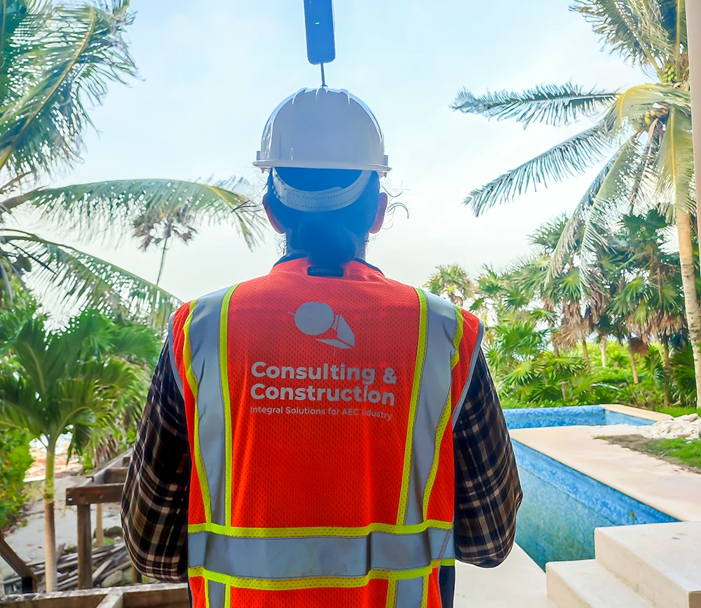A person wearing a white hard hat with a small camera and an orange safety vest stands among palm trees under a blue sky, likely capturing site conditions for a BIM Model workflow.