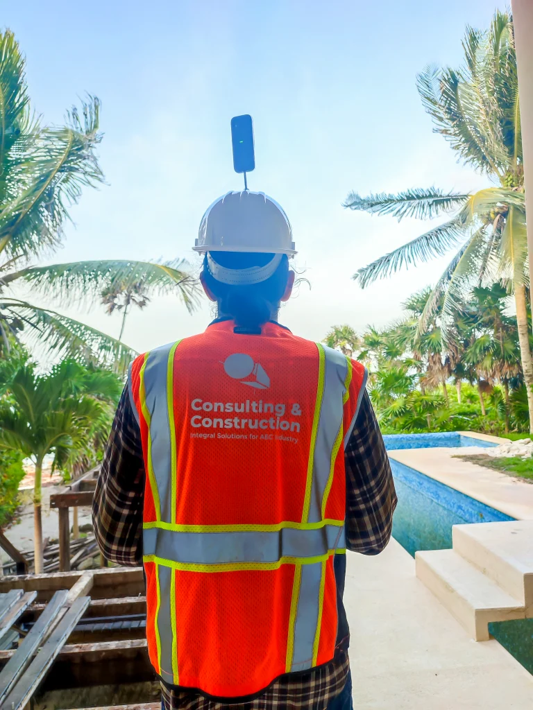 Una persona con un casco blanco con un dispositivo montado y un chaleco de seguridad naranja de Consulting & Construction junto a una piscina, frente a palmeras y vegetación tropical, demuestra el uso de una cámara 360 para la gestión de obras
