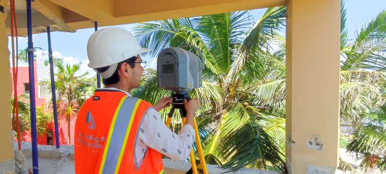 Un trabajador de la construcción con casco blanco y chaleco naranja maneja un instrumento topográfico sobre un trípode en el interior de un edificio en construcción, con palmeras visibles en el exterior