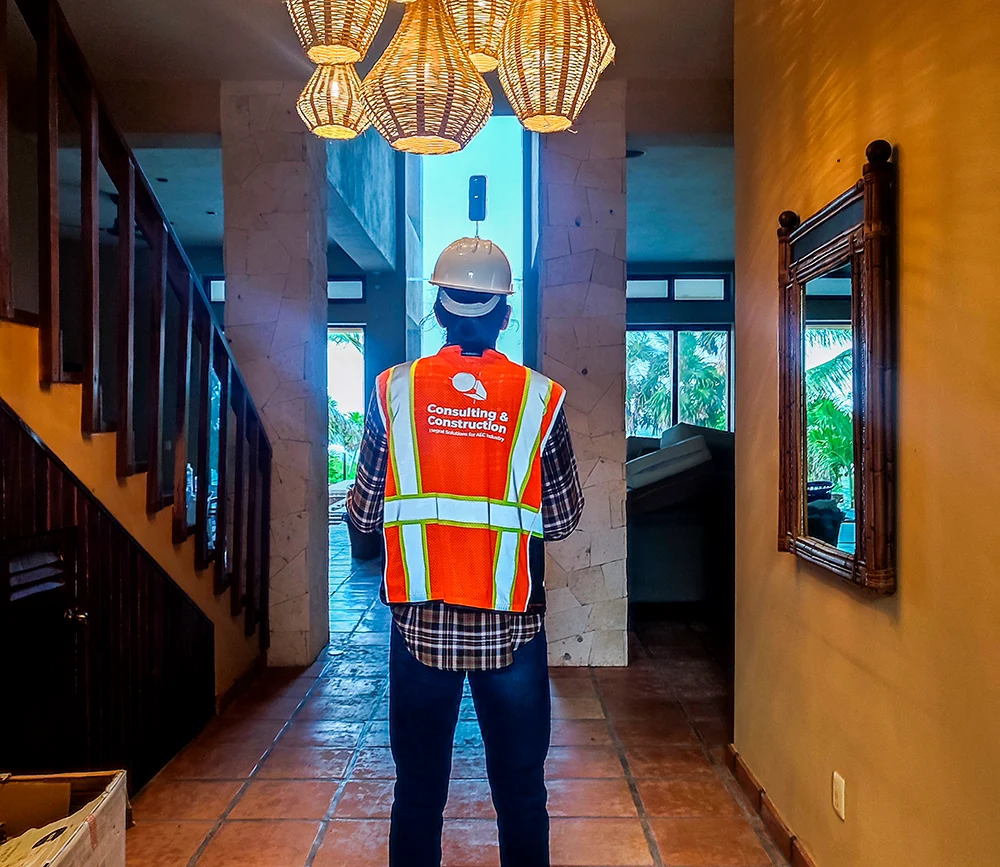A construction worker wearing a hard hat and orange safety vest stands inside, using a 360-degree camera on a pole to capture the space for the BIM model. The space features modern decor, large windows, and woven pendant lights overhead.