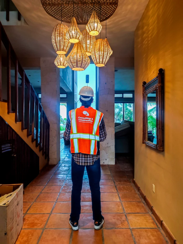 Una persona con casco y chaleco reflectante para la construcción se encuentra en un pasillo embaldosado con luces decorativas colgantes, de espaldas a la cámara hacia una ventana luminosa