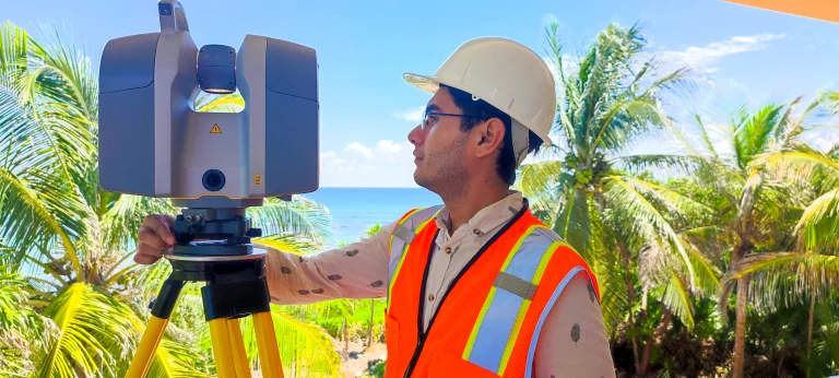 Un hombre con casco y chaleco de seguridad naranja maneja un instrumento topográfico sobre un trípode, capturando datos para un proyecto con palmeras y el océano de fondo bajo un cielo azul despejado