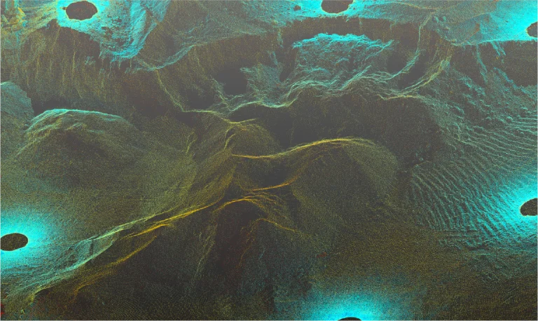 Vista aérea de un paisaje escarpado y montañoso con profundos valles y crestas, acentuado por puntos brillantes de color azul turquesa esparcidos por el terreno. La imagen tiene un efecto digital texturizado de Planta Industrial Nissan