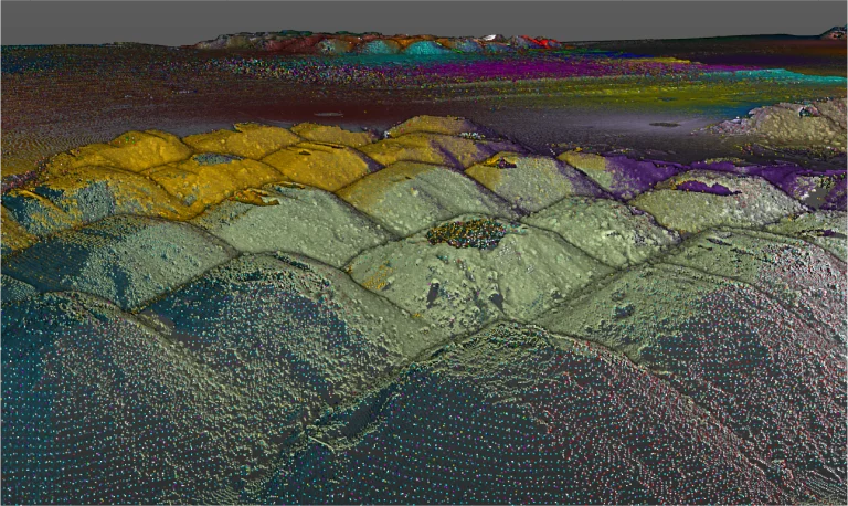 Un modelo digital del terreno renderizado en 3D de Planta Industrial Nissan, muestra hileras de montículos de color amarillo, verde, morado y rojo con una textura rugosa bajo un cielo nublado