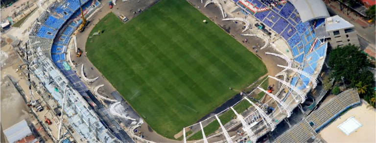 Vista aérea del Estadio Pascual Guerrero con un campo verde, asientos azules y blancos y obras visibles en un lateral. Se aprecian algunos equipamientos y zonas inacabadas en los bordes