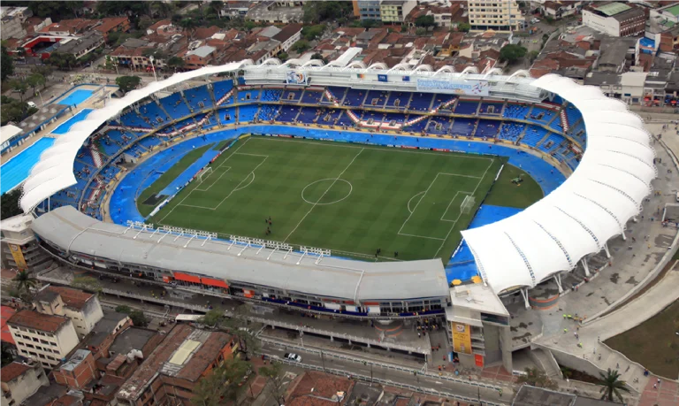 Vista aérea del Estadio Pascual Guerrero, un gran estadio de fútbol con un campo verde, asientos azules y un tejado curvo blanco sobre parte de las gradas, rodeado de edificios y carreteras de la ciudad