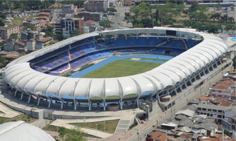 Vista aérea del Estadio Pascual Guerrero, con una pista de atletismo azul que rodea un campo de fútbol verde, asientos parcialmente cubiertos y edificios urbanos alrededor del recinto