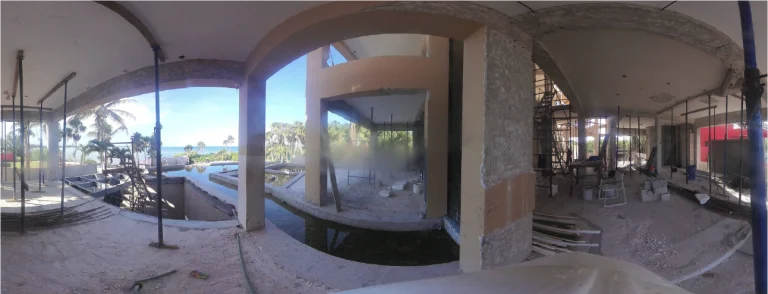 Vista panorámica de un edificio parcialmente construido con paredes abiertas, materiales de construcción dispersos, andamios y un atisbo de palmeras y el océano en el exterior