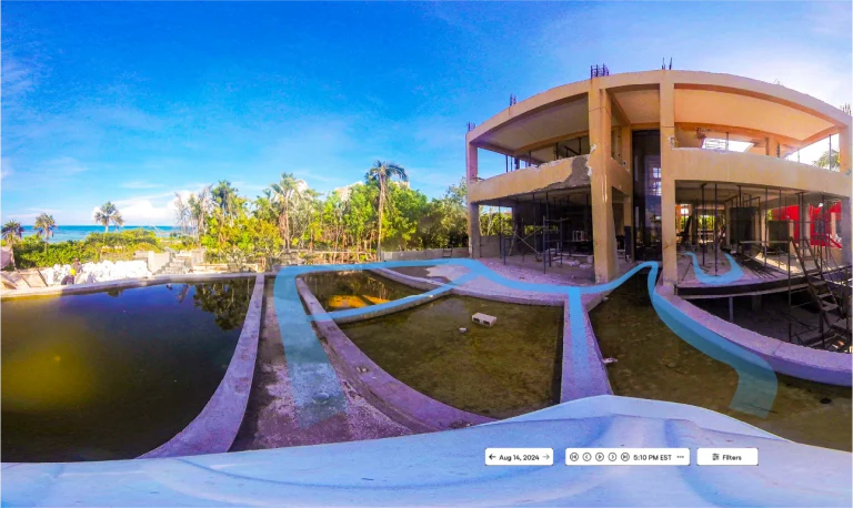 Una vista en gran angular de un edificio abandonado e inacabado con hormigón visto y piscinas vacías, rodeado de vegetación y palmeras,