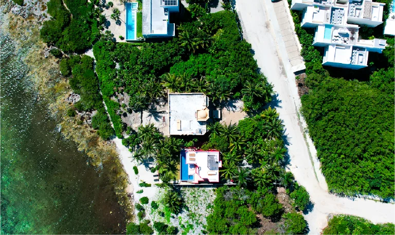 Vista aérea de casas modernas, rodeadas de densos árboles verdes cerca de una costa, con una carretera paralela al agua y una piscina visible en una propiedad