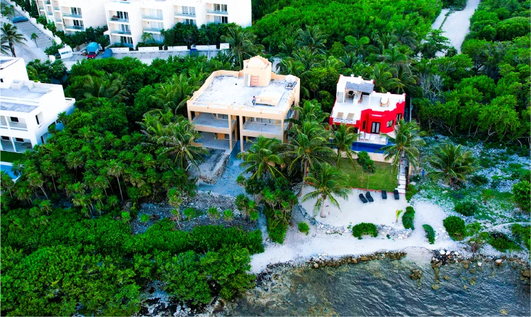 Vista aérea de dos casas junto a la costa, rodeadas de frondosos árboles. La casa beige con tejado inacabado podría beneficiarse de un Modelo BIM para agilizar su construcción en medio de las impresionantes aguas cristalinas y el primer plano rocoso