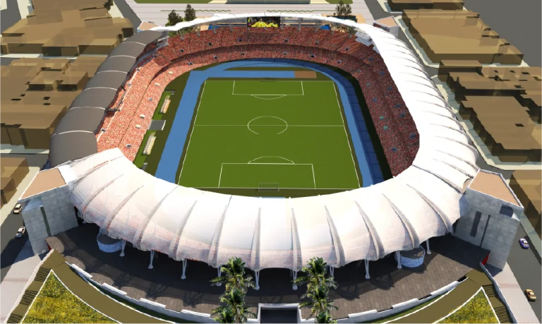 Vista aérea del Estadio Pascual Guerrero, un gran y moderno estadio de fútbol lleno de espectadores. El estadio presenta un techo blanco parcialmente cubierto y un campo de fútbol verde estándar, con edificios cercanos y carreteras visibles fuera del estadio