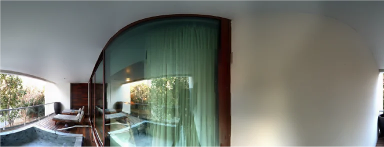 Una vista panorámica de un moderno balcón con tumbonas, una barandilla de cristal, una puerta de cristal curvada con cortinas y una pequeña piscina o bañera de hidromasaje: en el exterior se ven árboles, captados con todo detalle gracias a la tecnología Escaneo Láser Vidanta