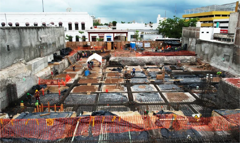 Una gran obra con obreros construyendo los cimientos de una nueva estructura. Muros de contención de hormigón, redes de seguridad naranjas y materiales llenan la obra, con edificios y árboles al fondo