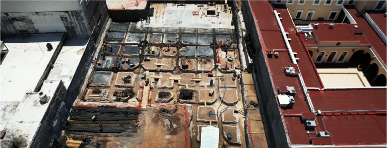 Vista aérea de una excavación rodeada de edificios, donde se ven trabajadores y materiales entre fosas rectangulares