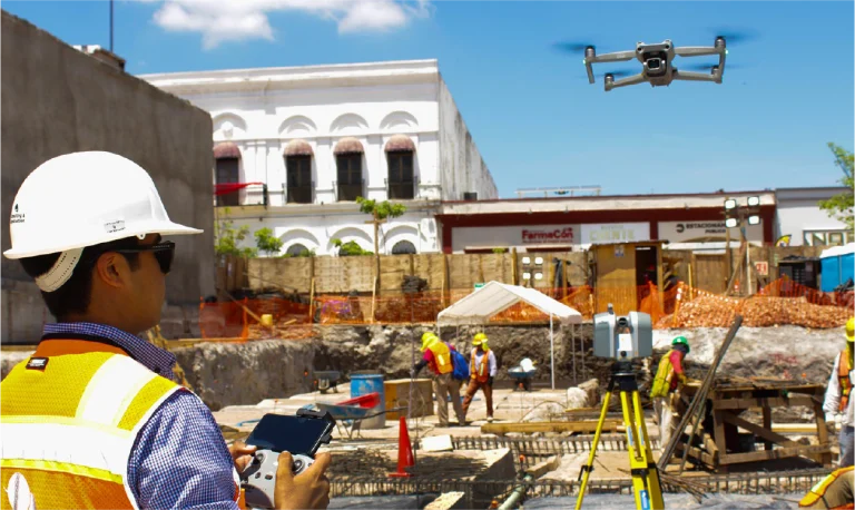 Un trabajador de la construcción con casco y chaleco de seguridad opera un dron para Escaneo Láser Coppel Culiacán en una obra urbana. Otros trabajadores y materiales de construcción son visibles en el fondo bajo un cielo brillante y soleado