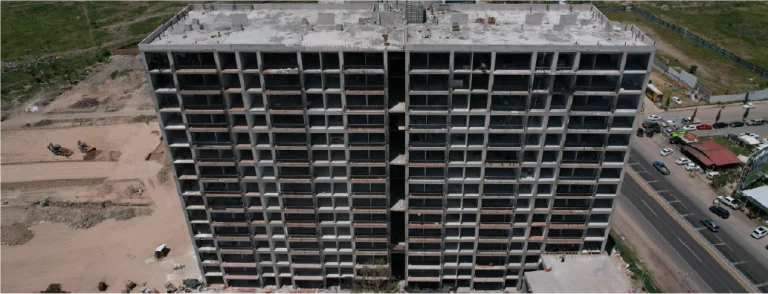 Un alto e inacabado edificio de apartamentos de hormigón del Gran Valbuena desarrollo de vivienda se alza junto a una carretera, rodeado de tierra y maquinaria de construcción, con ventanas abiertas y balcones a la vista en cada planta