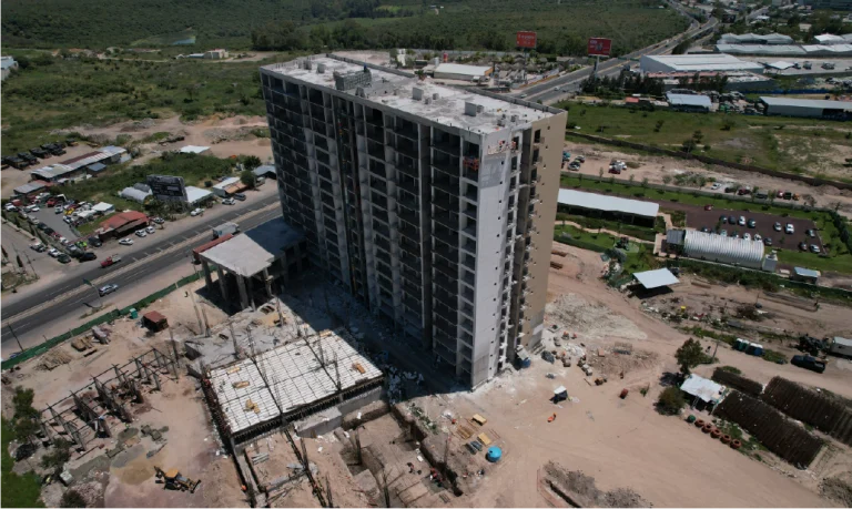 Vista aérea del desarrollo de viviendas Grand Valbuena, que muestra un edificio alto en construcción, rodeado de materiales y equipos, con carreteras, vegetación y otros edificios al fondo