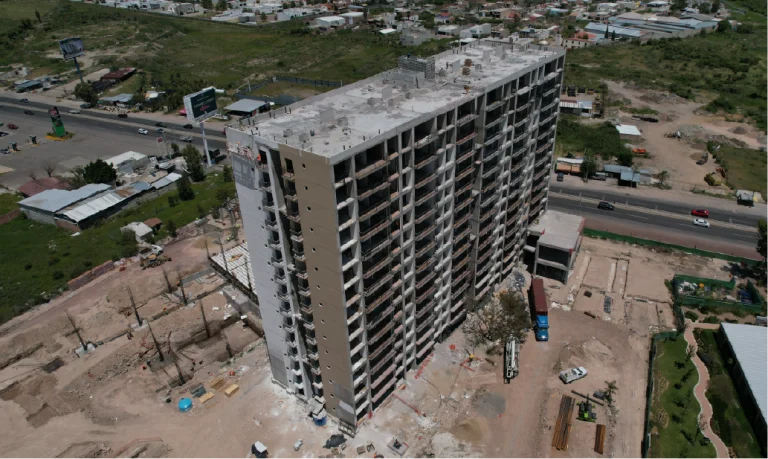 Vista aérea de Grand Valbuena, una vivienda alta e inacabada rodeada de tierra, materiales de construcción, vehículos y una carretera principal con vegetación al fondo
