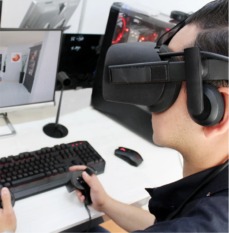 Una persona con un casco de realidad virtual y auriculares utiliza unos controladores de mano en un escritorio con teclado, ratón y monitor que muestra una vivienda o urbanización virtual en Puerta de Hierro