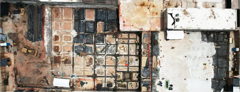 Vista aérea de una obra en construcción con trabajadores, maquinaria y secciones de tierra y hormigón en forma de cuadrícula. Las zonas adyacentes muestran edificios y tejados blancos