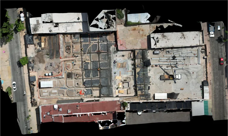Vista aérea de un sitio de construcción en Escaneo Láser Coppel Culiacán, rodeado de edificios y carreteras, mostrando cimientos expuestos, materiales de construcción, vehículos y trabajadores dentro de una manzana rectangular