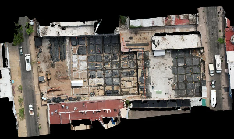 Vista aérea de una obra en construcción en una zona urbana, que muestra una cuadrícula de secciones excavadas, materiales de construcción, trabajadores y maquinaria