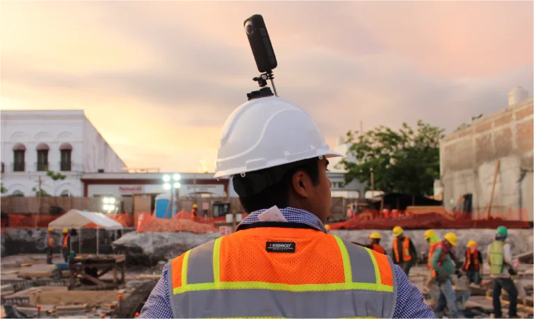 Un trabajador de la construcción con un casco blanco y un chaleco de seguridad naranja se encuentra en una obra al atardecer, mientras otros trabajadores con cascos trabajan al fondo