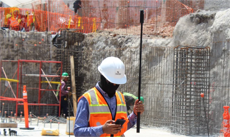 Un trabajador de la construcción con casco blanco, chaleco de seguridad naranja y cubierta facial sostiene un poste de medición y revisa su teléfono inteligente durante un proyecto de Escaneo Láser Coppel Culiacán en un sitio de construcción activo con marcos de barras de refuerzo y compañeros de trabajo en el fondo