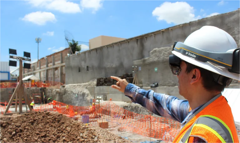 Un trabajador de la construcción con casco, chaleco de seguridad y gafas de sol señala hacia una obra con montones de tierra, redes de seguridad naranjas, muros de hormigón y trabajos en curso