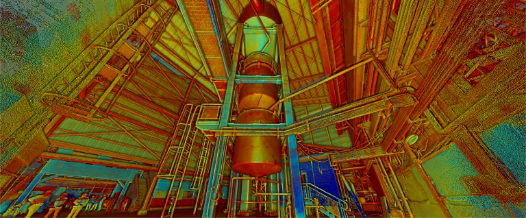 Un cohete de gran altura se alza verticalmente en el interior de un edificio de aspecto industrial en el Ingenio Azucarero Tamazula, en Jalisco, rodeado de vigas metálicas, tuberías y pasarelas. Varias personas contemplan el cohete en medio de fuertes tonos rojos y amarillos