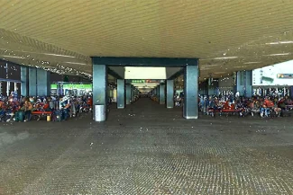 Un gran pasillo abierto del aeropuerto de Tijuana cuenta con hileras de bancos a ambos lados, donde muchas personas están sentadas bajo un techo a rayas. La espaciosa zona está flanqueada por columnas estructurales que se extienden en la distancia.