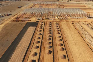 An aerial view of the BMW Industrial Plant construction site in a desert area, showing rows of evenly spaced pits, steel frame structures, and construction vehicles scattered around the site.