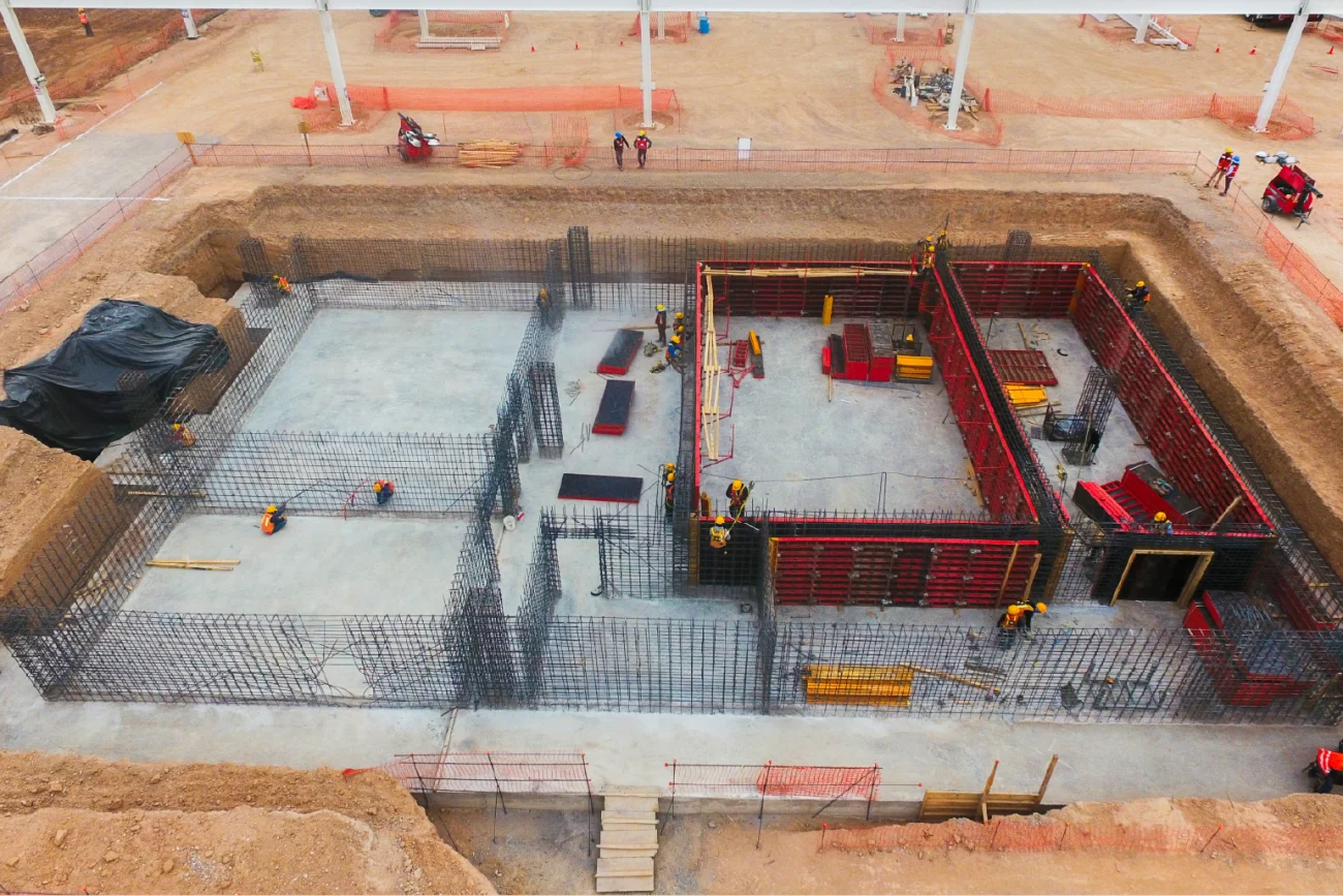 An aerial view of a construction site at the BMW Industrial Plant, with workers reinforcing concrete foundations and steel rebar inside a large rectangular excavation surrounded by earth and safety barriers.