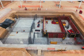 An aerial view of a construction site at the BMW Industrial Plant, with workers reinforcing concrete foundations and steel rebar inside a large rectangular excavation surrounded by earth and safety barriers.