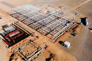 An aerial view of a construction site in a desert area, showing the large steel frame of a future BMW Industrial Plant, with construction equipment, workers, and foundations dug into sandy soil for the automotive industry.