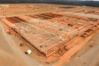 An aerial view of the BMW Industrial Plant construction site, showing steel structures rising above a vast expanse of cleared red earth. Several rows of metal beams and construction vehicles are visible on the site.