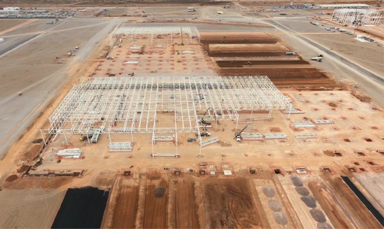 Vista aérea de una gran obra de construcción en una zona desértica, con estructuras de acero para la Planta Industrial BMW, vehículos de construcción y parcelas marcadas sobre suelo marrón rojizo, con carreteras y equipos visibles al fondo