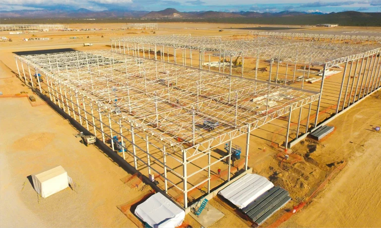 Vista aérea de una gran Planta Industrial BMW en construcción en una zona desértica, con armazón de acero elevándose, materiales de construcción y equipos esparcidos alrededor, y montañas visibles al fondo
