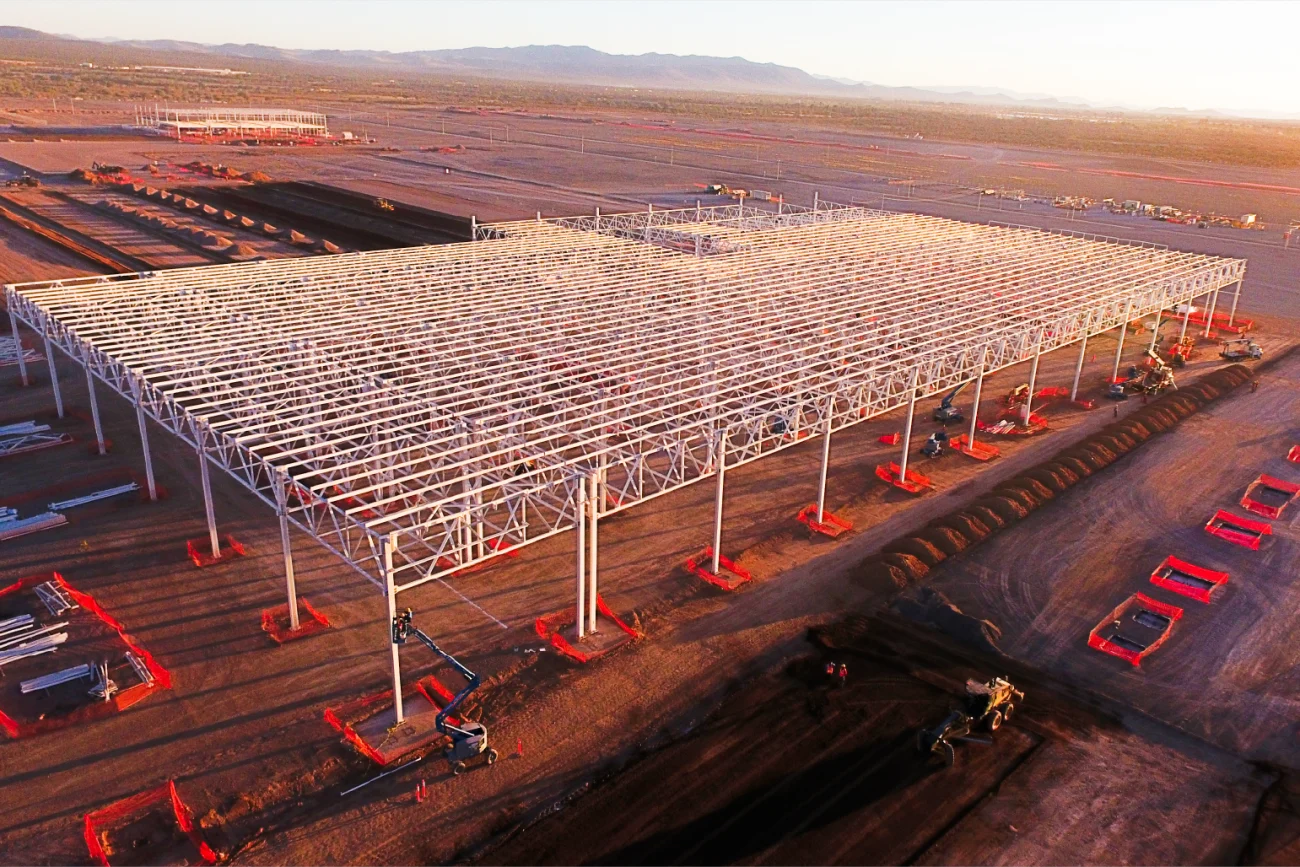 An aerial view of the BMW Industrial Plant construction site, featuring a steel structure under construction, surrounded by construction materials, machinery, and soil in an open landscape.