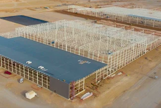 A large BMW Industrial Plant under construction in a desert area, with visible steelwork, partially installed roof panels, and various smaller structures and equipment around the site.