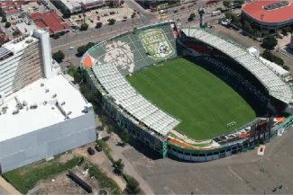 Aerial view of the Nou Camp Stadium, a large football stadium with green seats, surrounded by urban buildings and parking lots under a clear sky. The well-maintained pitch and stands feature striking designs and text.