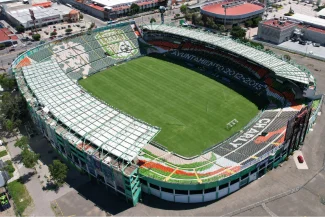 Aerial view of the Nou Camp Stadium, a large, empty football stadium with green stands and a well-maintained pitch, surrounded by buildings and streets.
