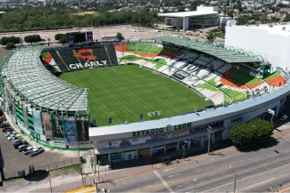 Aerial view of León Stadium, a large football stadium with green, orange, black, and white seats, a green field, and partially covered stands in an urban area with trees and nearby buildings.