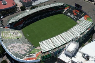 Aerial view of the Nou Camp stadium, with a green field, multicolored seats, and partially covered stands; CHARLY and CITY COUNCIL 2013-2015 are visible in the seats. Streets and buildings surround the stadium.