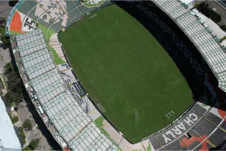 Aerial view of an empty soccer stadium with the green field, surrounding stands, and some covered seating areas. The word GRAND is visible in the lower right corner of the stands at Pascual Guerrero Stadium.