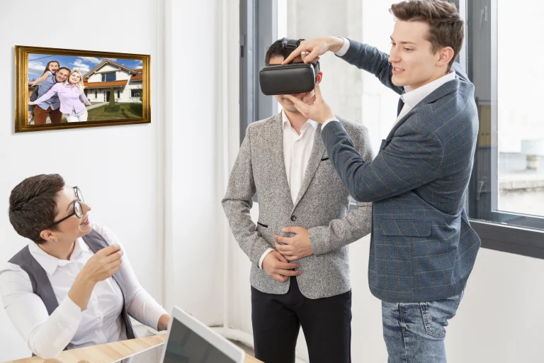 Tres personas en una oficina; un hombre ayuda a otro a ponerse unas gafas de realidad virtual para explorar la realidad extendida, mientras una mujer observa y sonríe. Una foto familiar cuelga en la pared detrás de ellos.