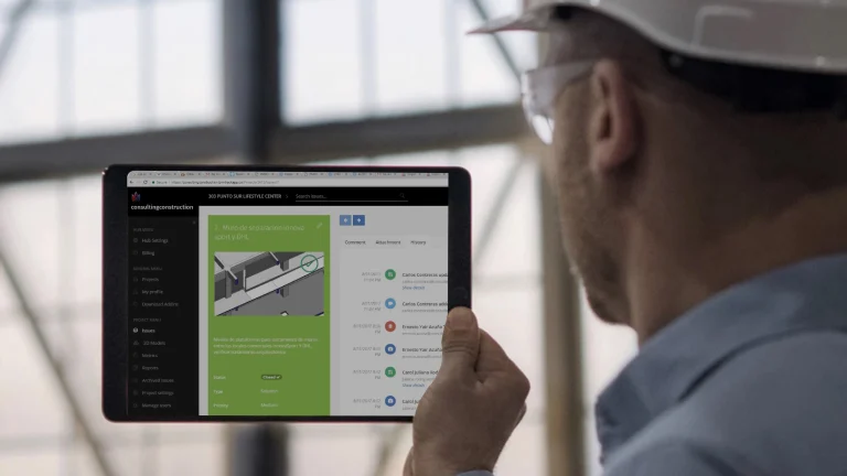 A person wearing a white safety helmet and glasses holds a tablet displaying modelos digitales with construction diagrams and text. The background reveals a blurred industrial setting with large windows.